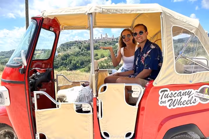 Smiling pair on Tuscany wine tour by tuk-tuk with stunning countryside backdrop and medieval town in view.