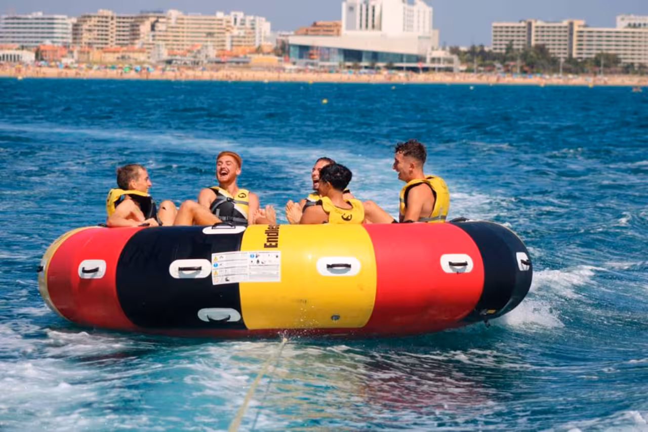 Excited group riding Twister inflatable off resort coastline, gripping handles as speedboat tows them through bright blue water