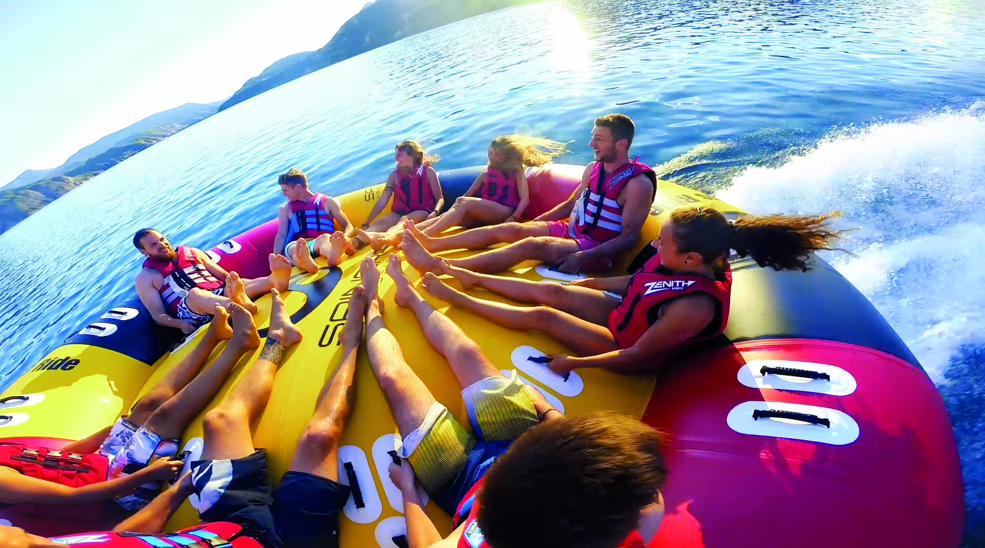 Group of adults enjoying thrilling Twister towable tube ride on sunny open water with life vests on.