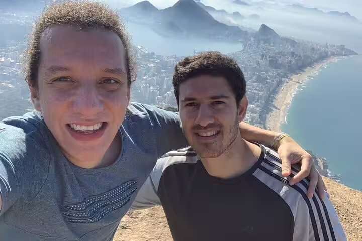 Two hikers smile for a selfie with a stunning backdrop of Rio de Janeiro's coastline from Two Brothers Hill.