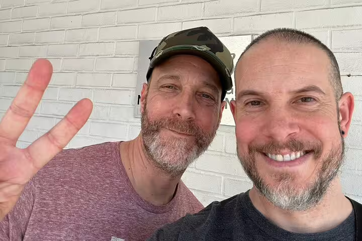 Two smiling men with beards taking a selfie against a white brick wall outdoors