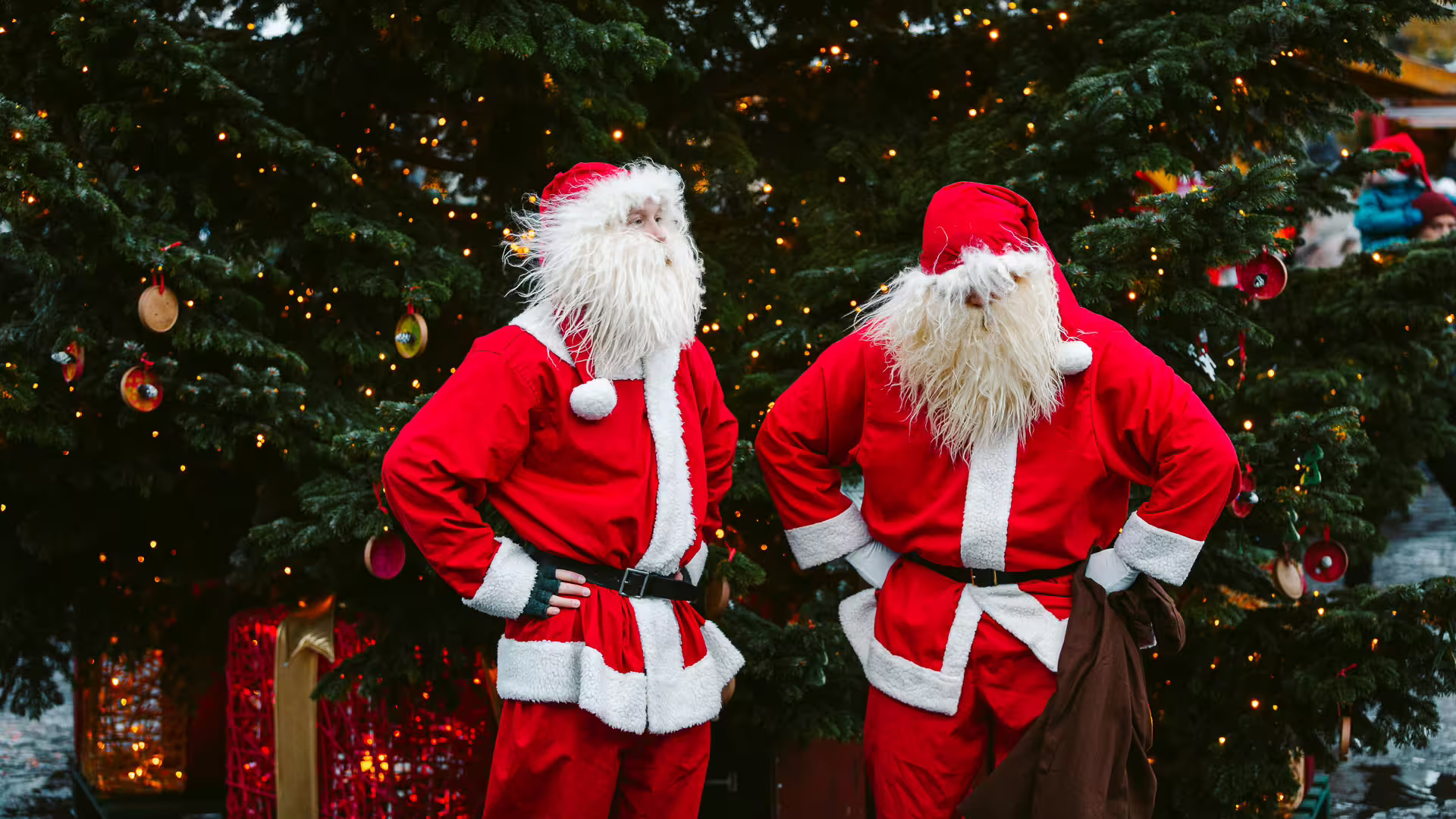 Two people dressed as Santa Claus standing in front of decorated Christmas trees with lights and ornaments
