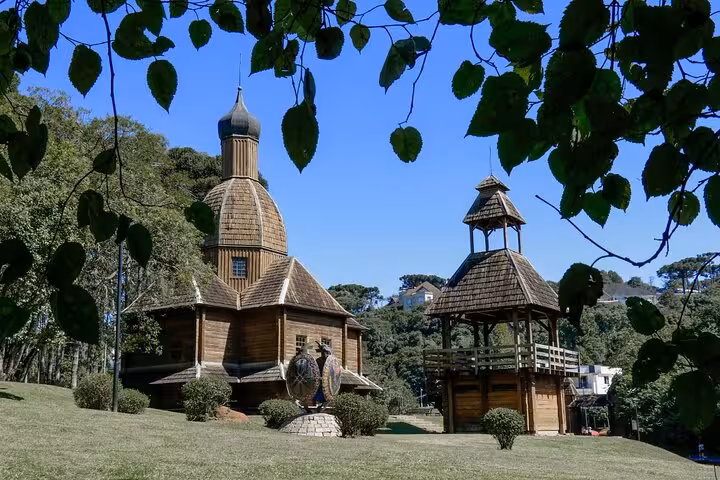 Charming wooden Ukrainian church and bell tower in Curitiba's Tingui Park, surrounded by lush greenery under a clear blue sky.