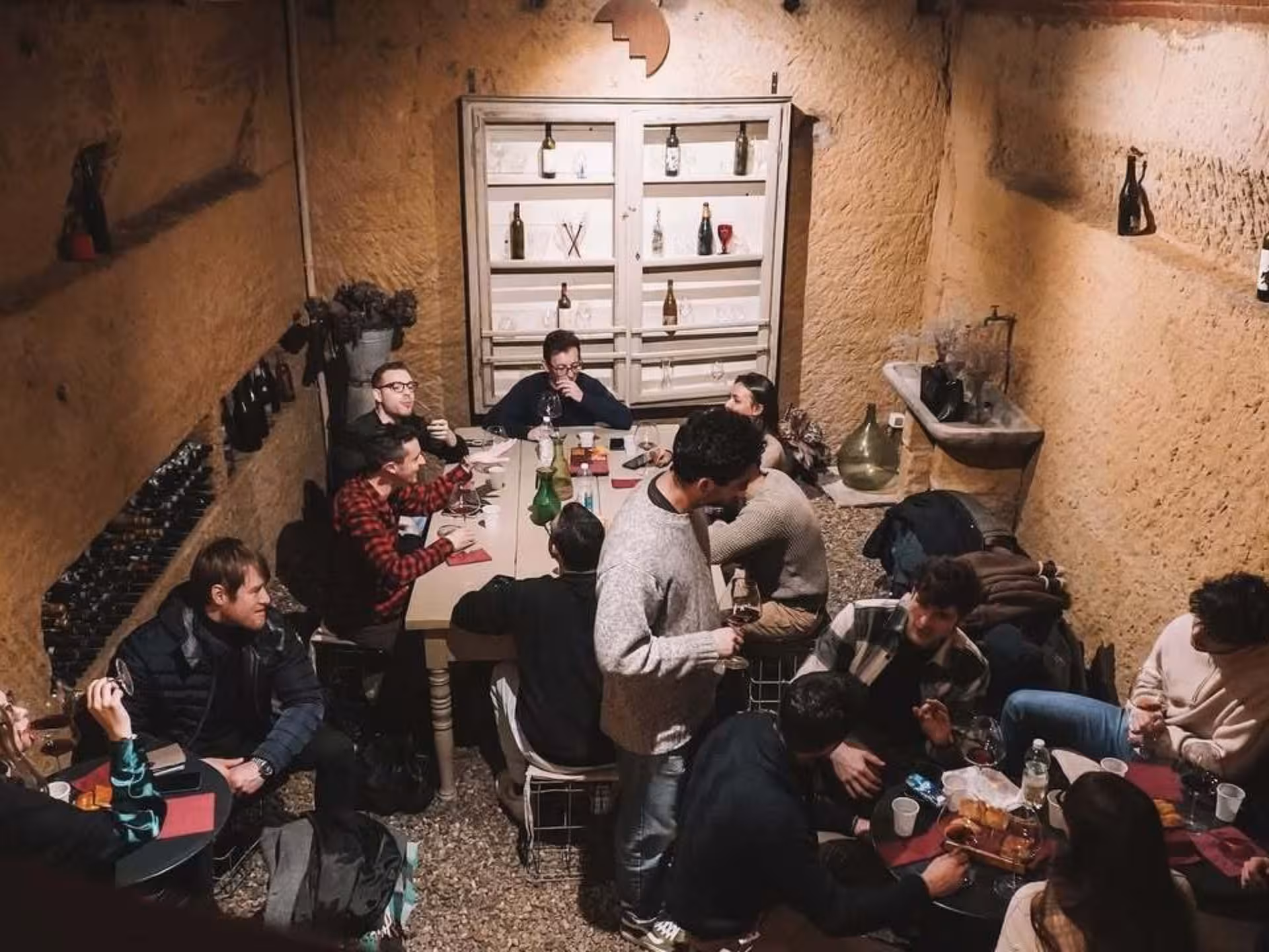 Group enjoying wine tasting at a rustic underground cave near Asti, surrounded by bottles and cozy decor.