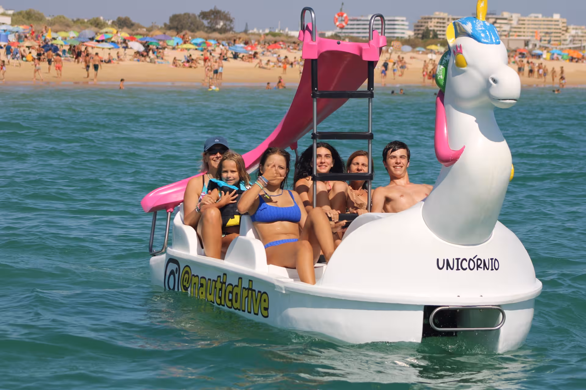 Happy family riding a unicorn-shaped pedal boat with a slide near a sunny beach on a kids and family tour.