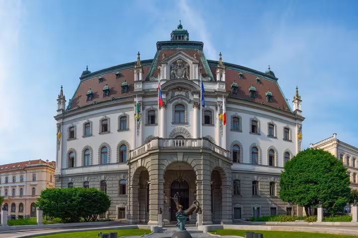 Grand neoclassical University of Ljubljana building with green dome, flags, and fountain in foreground