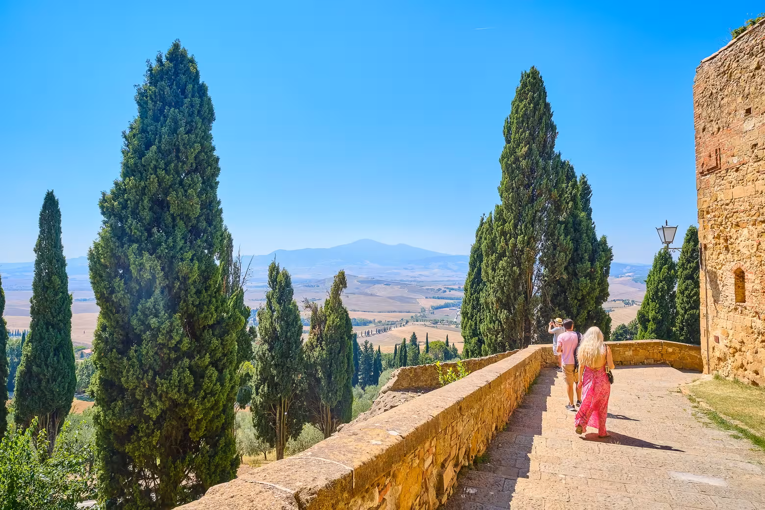 Tourists stroll along a scenic pathway in Val D'Orcia, surrounded by tall cypress trees and rolling Tuscan hills.
