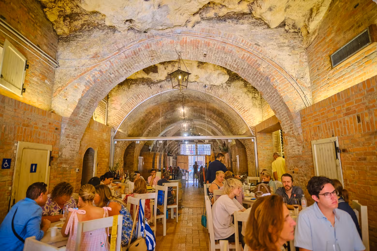 Visitors enjoy wine and cheese tasting in a rustic brick cellar in Val D'Orcia, featuring arched ceilings and warm lighting.