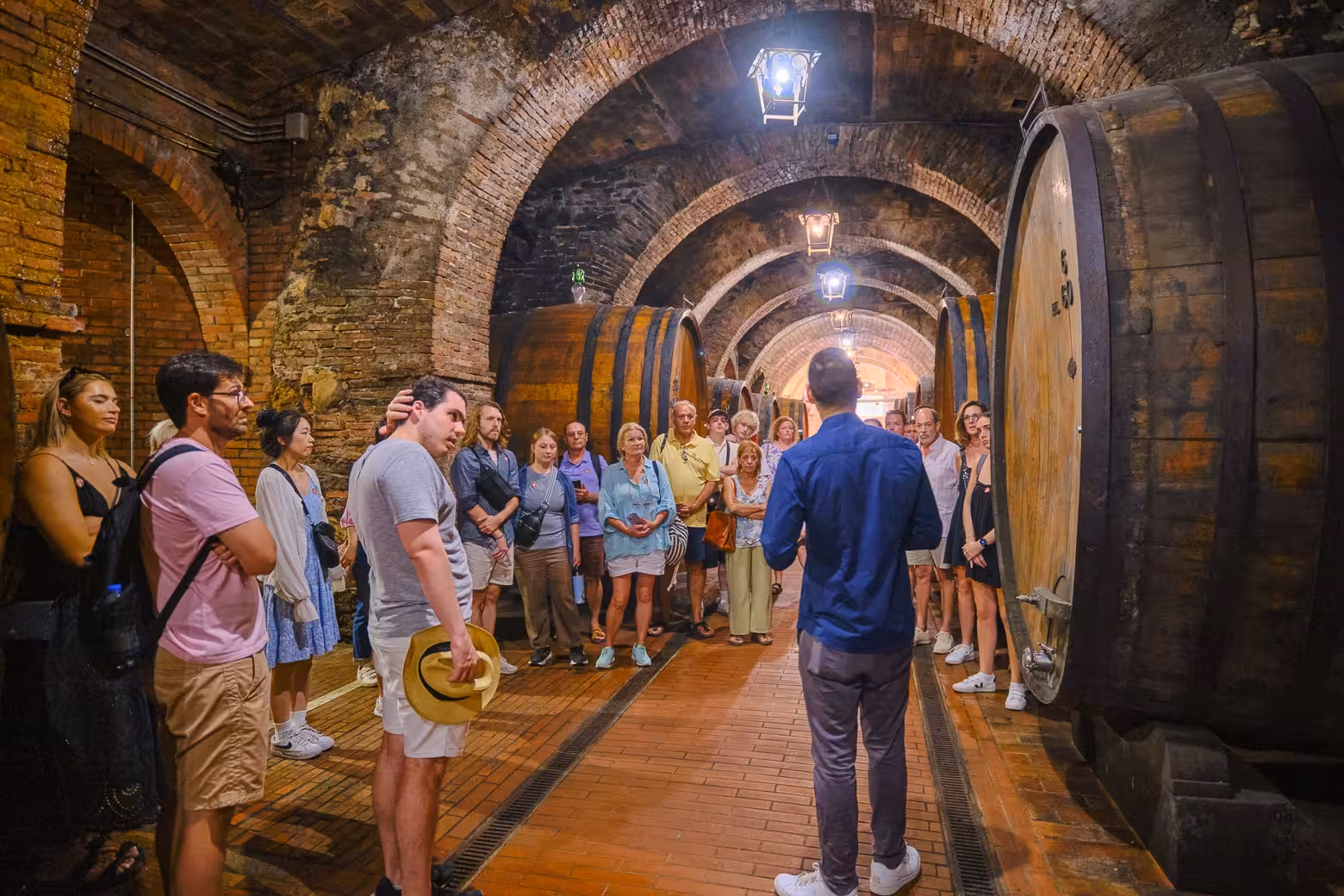 Guide leading a wine cellar tour in Val D'Orcia, surrounded by large wooden barrels and attentive visitors.