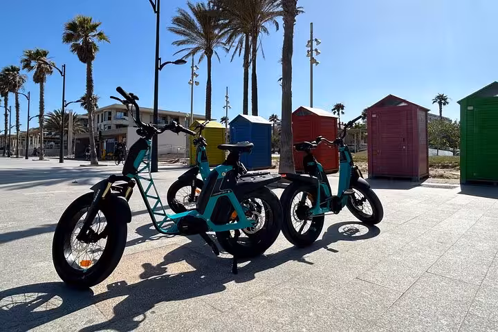 E-bikes parked by palm-lined Valencia beach promenade near colorful cabins on coastal tour stop
