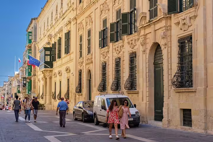 Tourists walking along a historic limestone street in Valletta, Malta, with ornate baroque buildings and green shutters