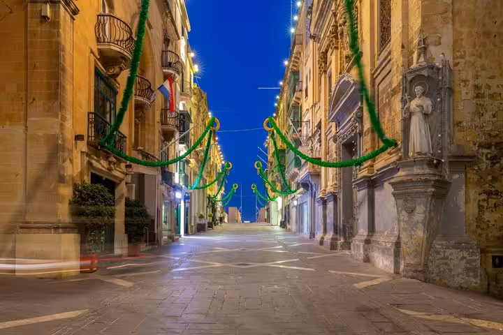 Valletta pedestrian street decorated with green festive garlands at dusk, historic limestone buildings lining both sides
