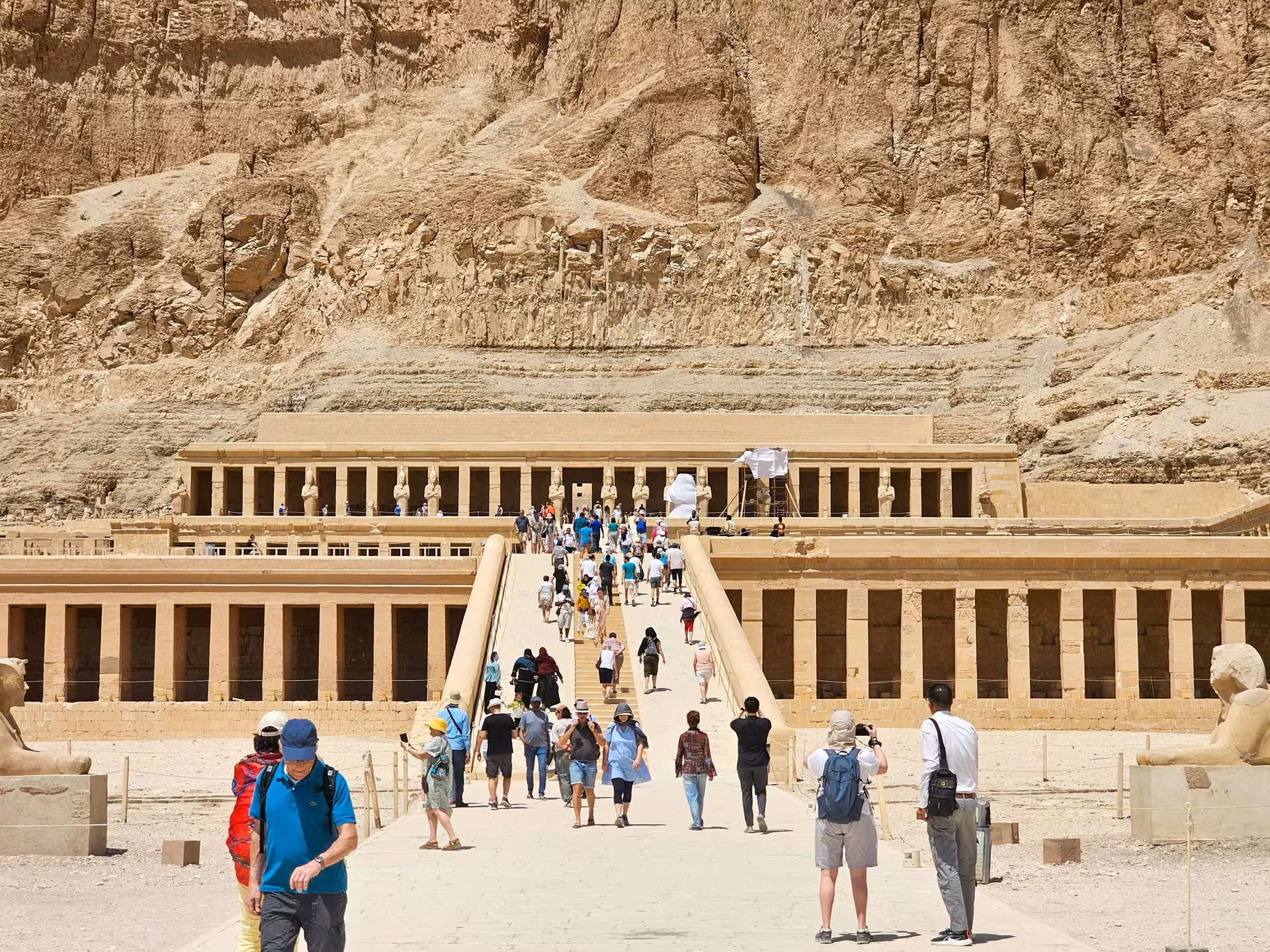 Tourists walking toward Hatshepsut Temple colonnades with towering limestone cliffs in Luxor, Egypt
