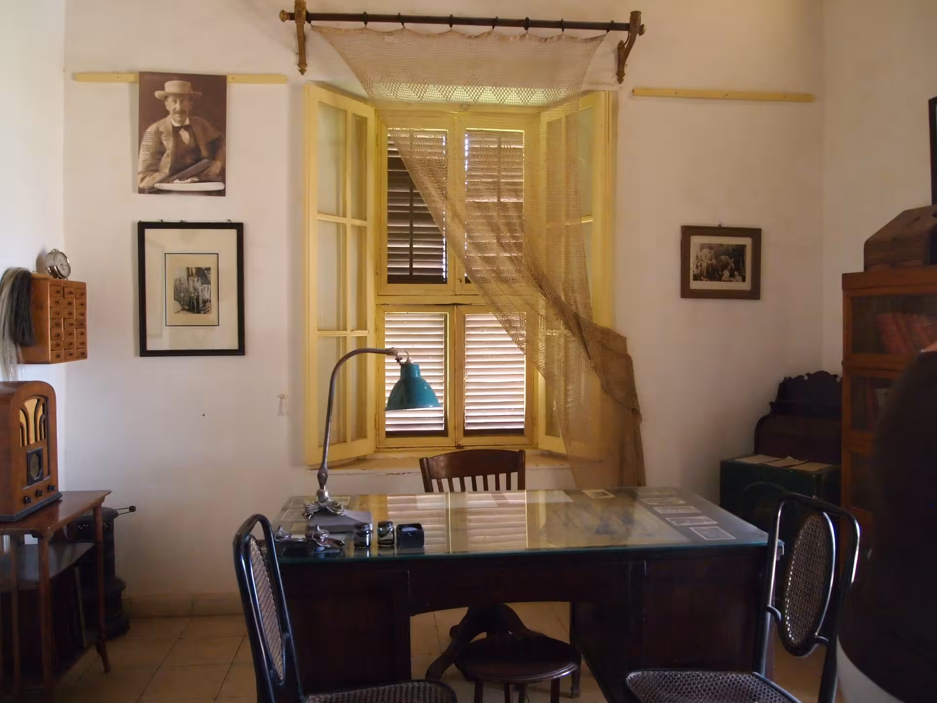 Historic office room with glass-top desk, vintage radio, open yellow shuttered window, and sepia portrait on wall
