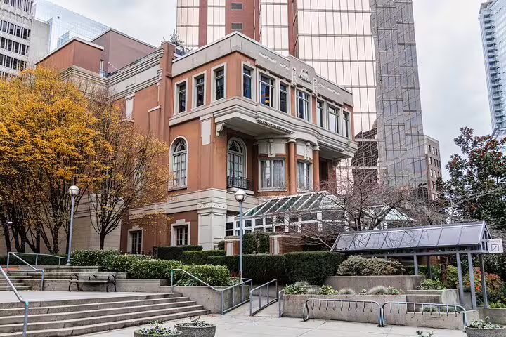 Historic brick mansion beside modern glass skyscraper in Vancouver, surrounded by autumn trees and manicured hedges