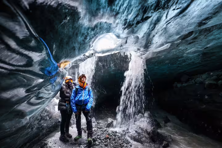 Two guided tour visitors stand beneath cascading ice waterfall inside stunning Vatnajokull crystal ice cave