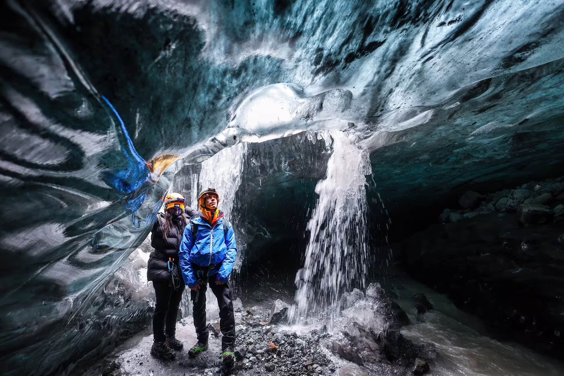 Two guided tour visitors stand beneath cascading ice waterfall inside stunning Vatnajokull crystal ice cave