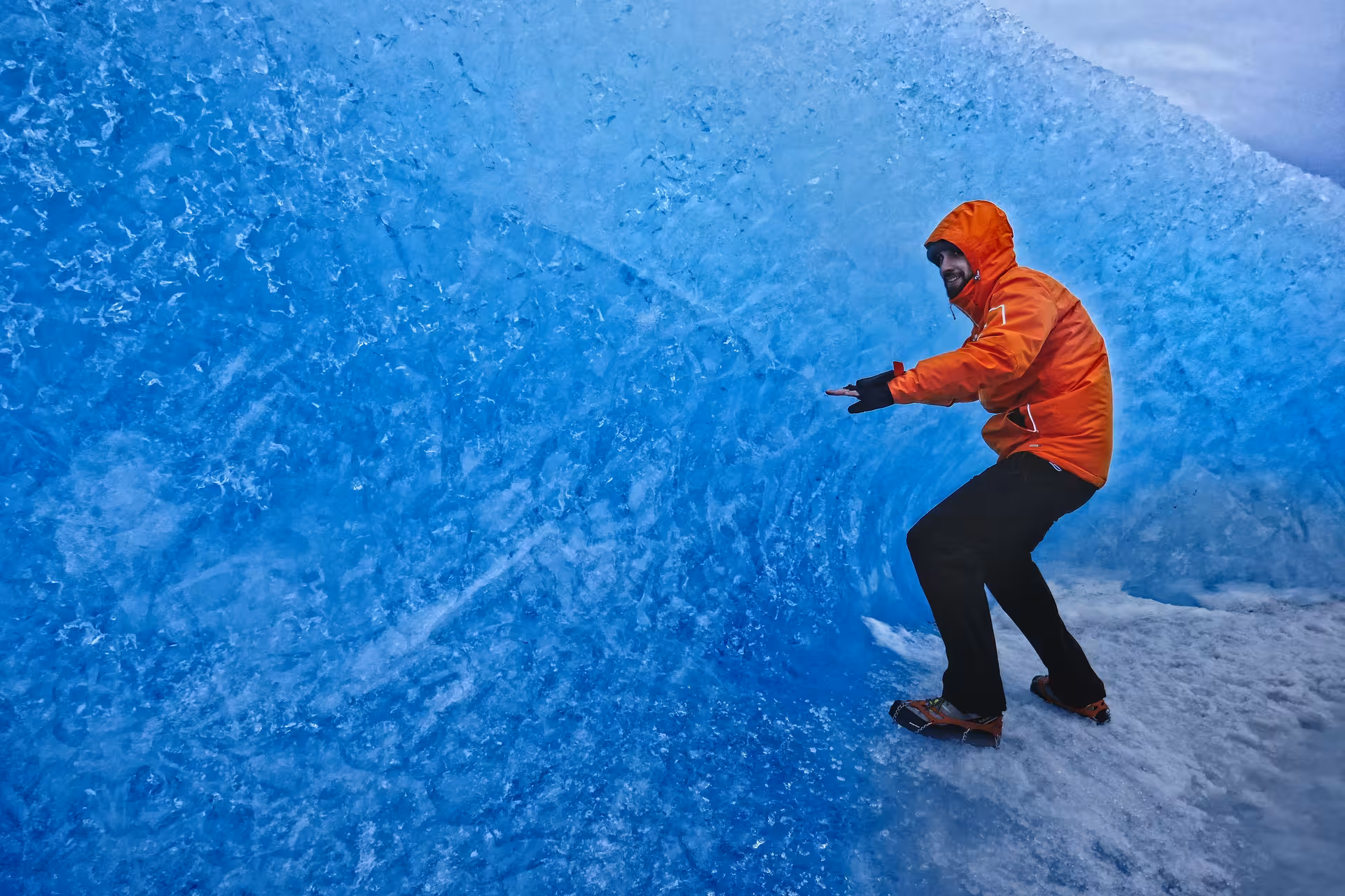 Explorer in orange jacket posing inside blue Vatnajokull glacier ice cave with crampons