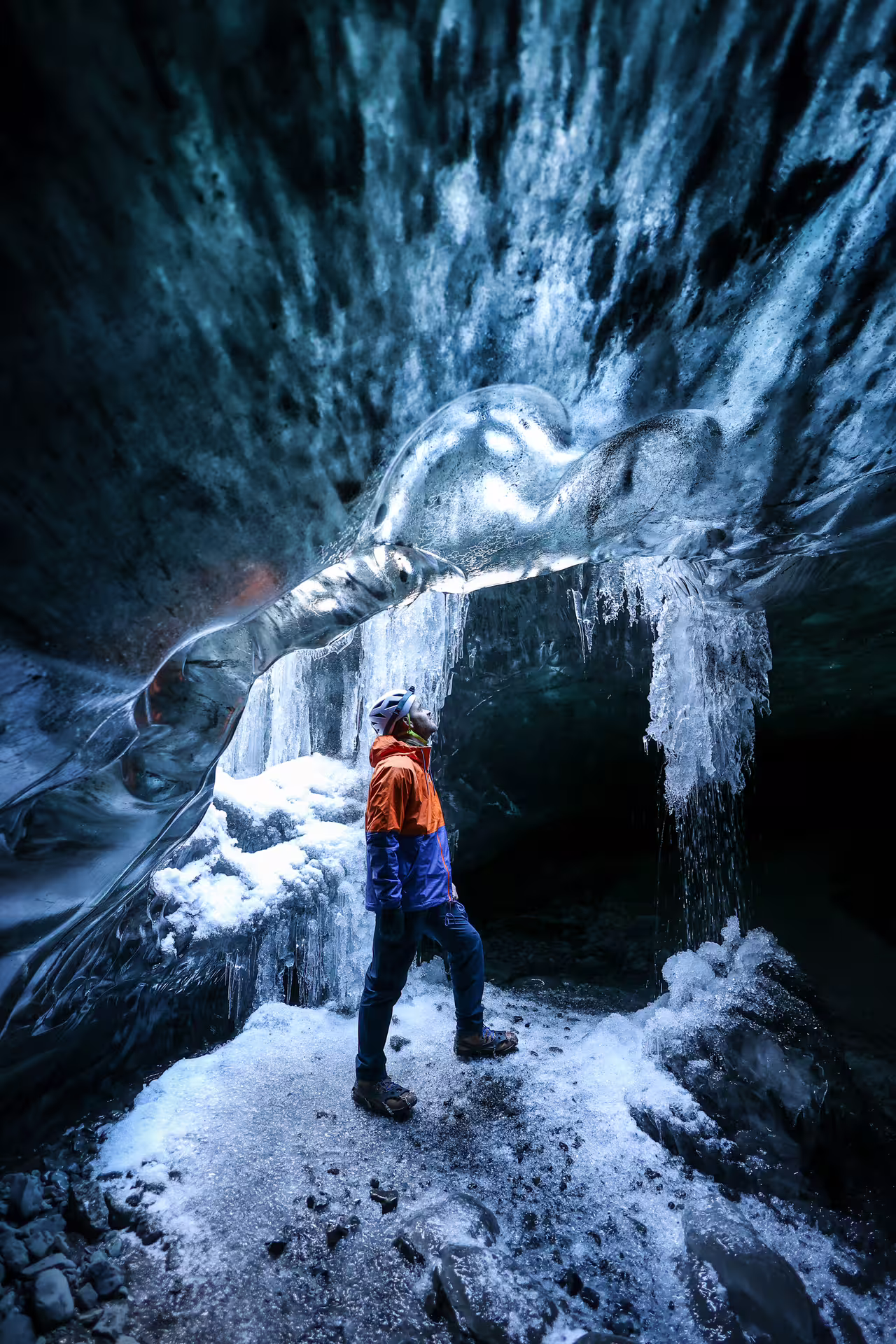 A solo explorer in an orange jacket gazes up at a glowing ice arch inside a stunning Vatnajokull Glacier ice cave.