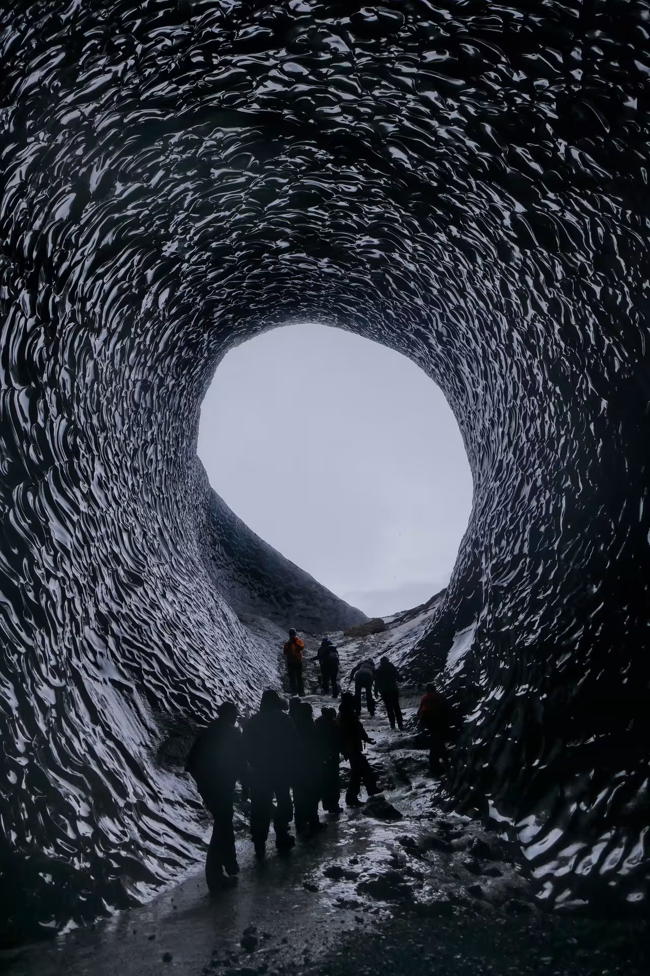 Tour group silhouetted inside a dark glacial ice cave tunnel with circular opening at Vatnajokull, Iceland