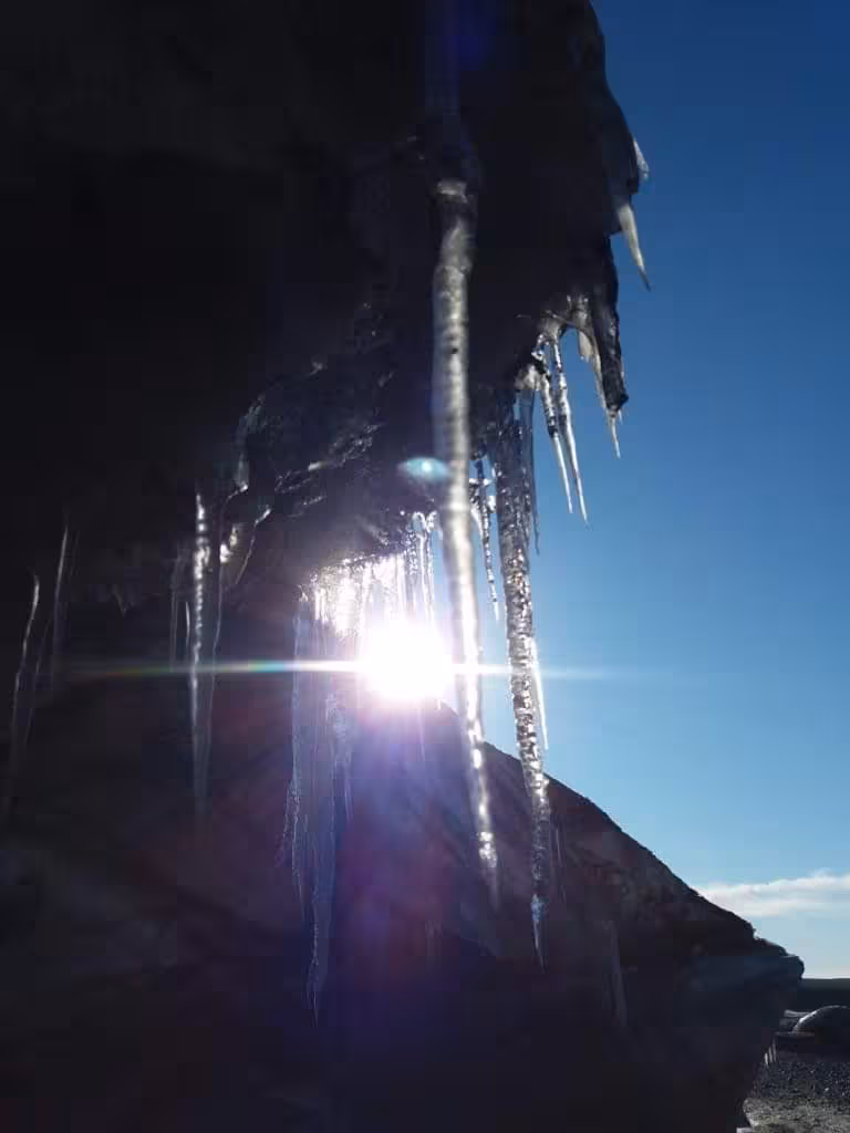 Icicles hanging from dark volcanic rock with sun flare and blue sky at Vatnajokull glacier