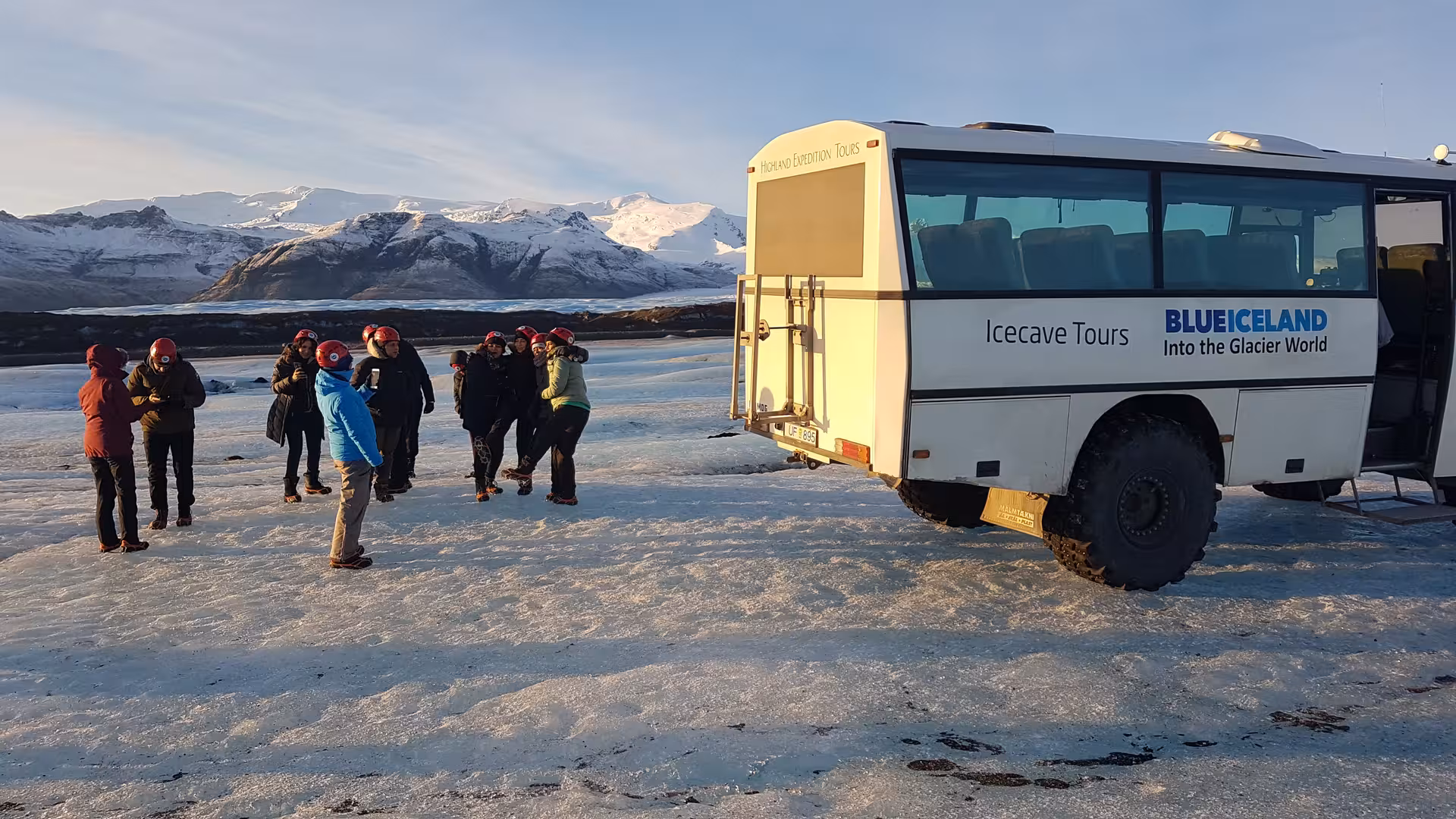 Tour group in red helmets beside Blue Iceland ice cave tour bus on Vatnajokull glacier with snowy mountains