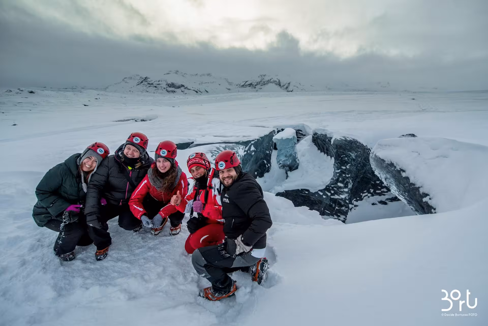 Group of five tourists in red helmets and crampons posing on Vatnajokull glacier near ice cave opening