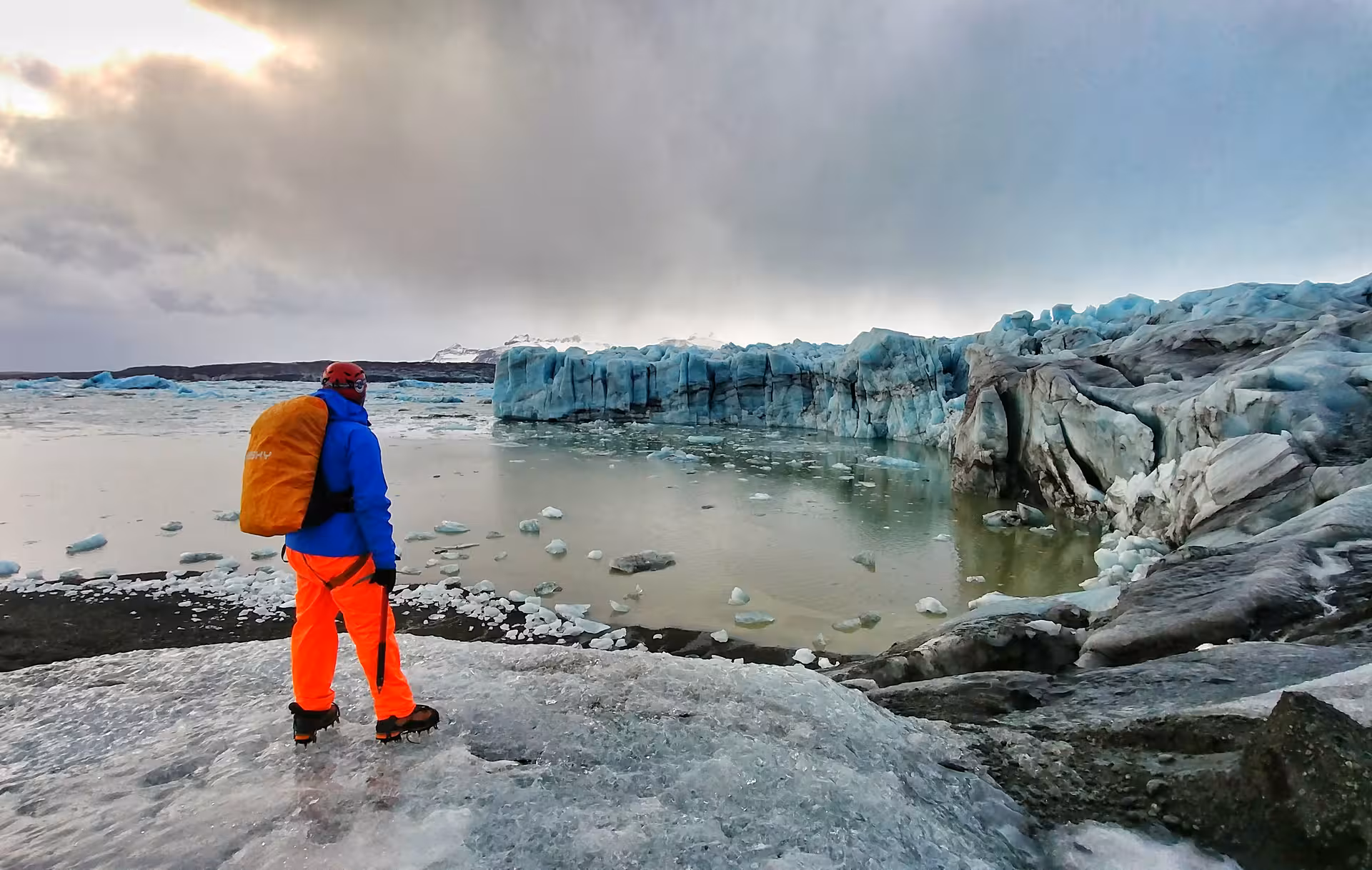 Hiker in orange pants and blue jacket standing on Vatnajokull glacier edge overlooking icy lagoon in Iceland