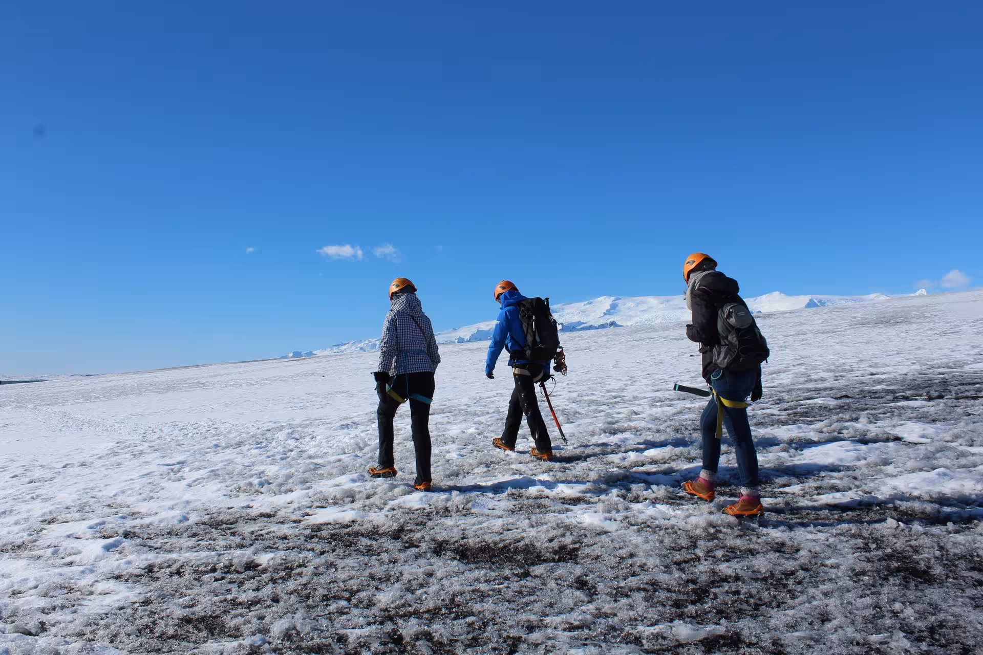 Three hikers with orange helmets and crampons walking on Vatnajokull glacier under clear blue sky
