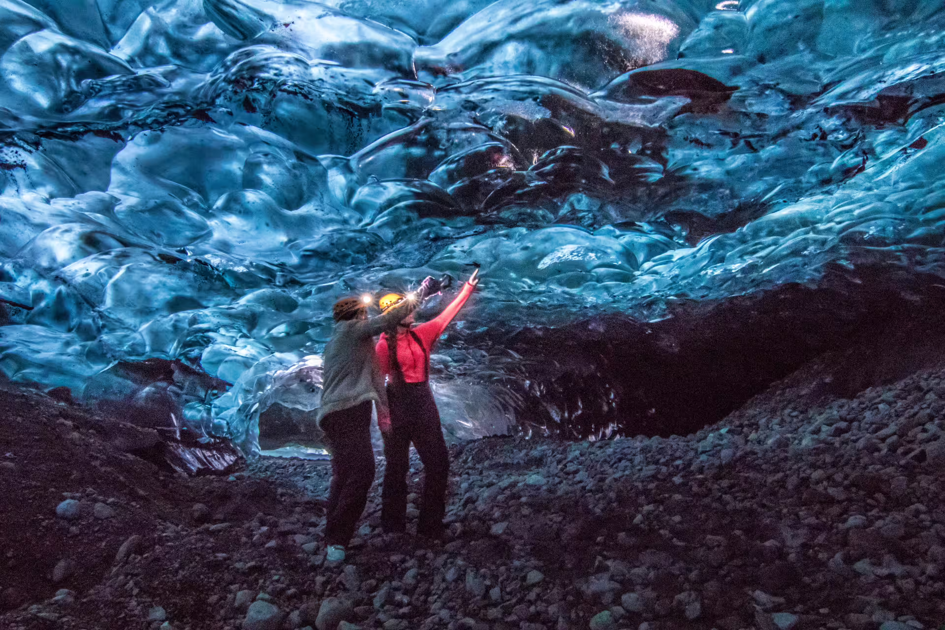 Two explorers with headlamps pointing at glowing blue ice cave ceiling in Vatnajokull glacier, Iceland