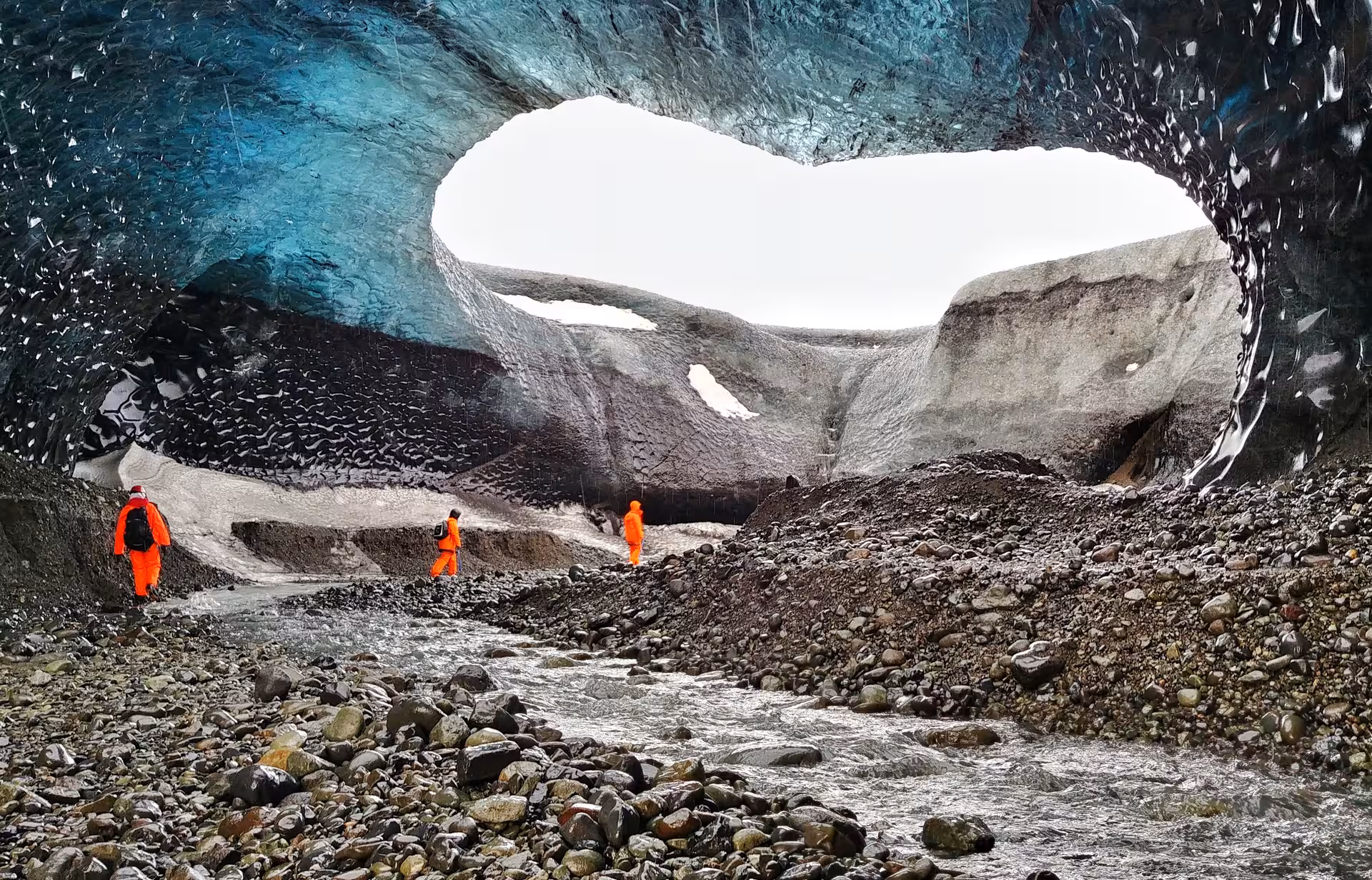 Three explorers in orange suits inside Vatnajokull glacier ice cave with blue ice ceiling and rocky stream