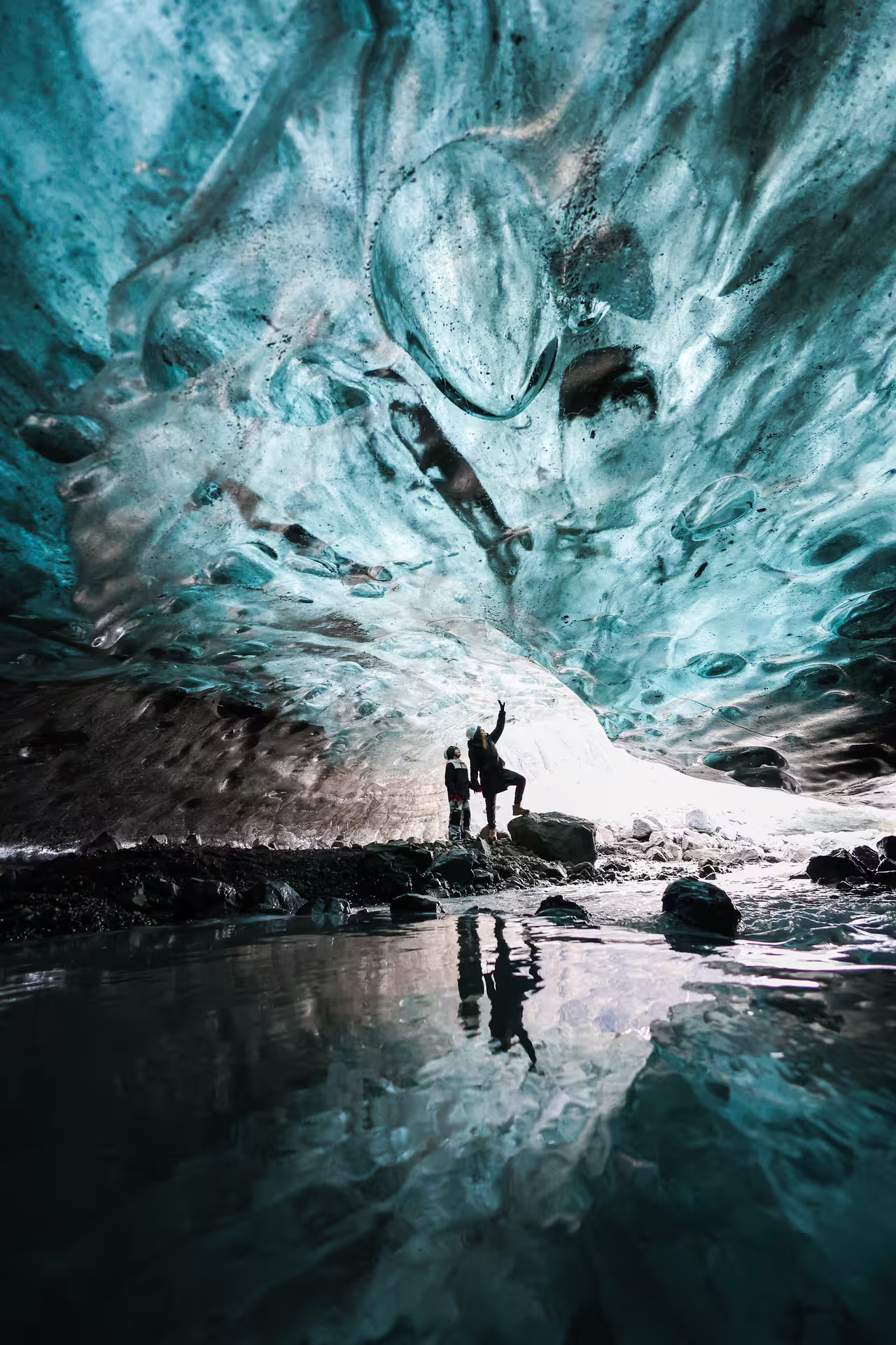 Two explorers reflected in water admiring vivid turquoise glacial ice cave walls on Eggtrip Winter Ice Cave tour Iceland
