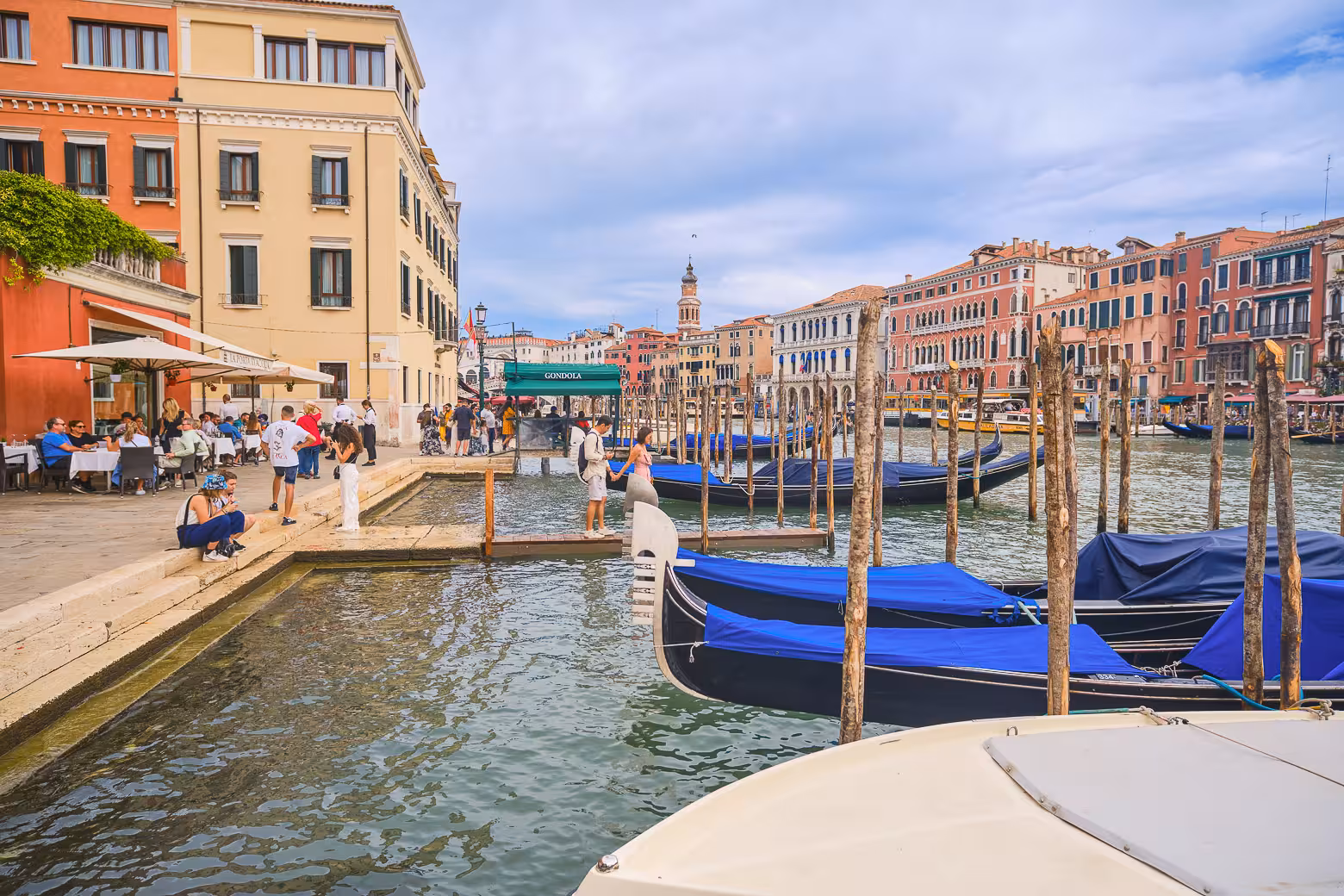 Charming Venice canal scene with gondolas docked beside colorful historic buildings and bustling outdoor cafes.