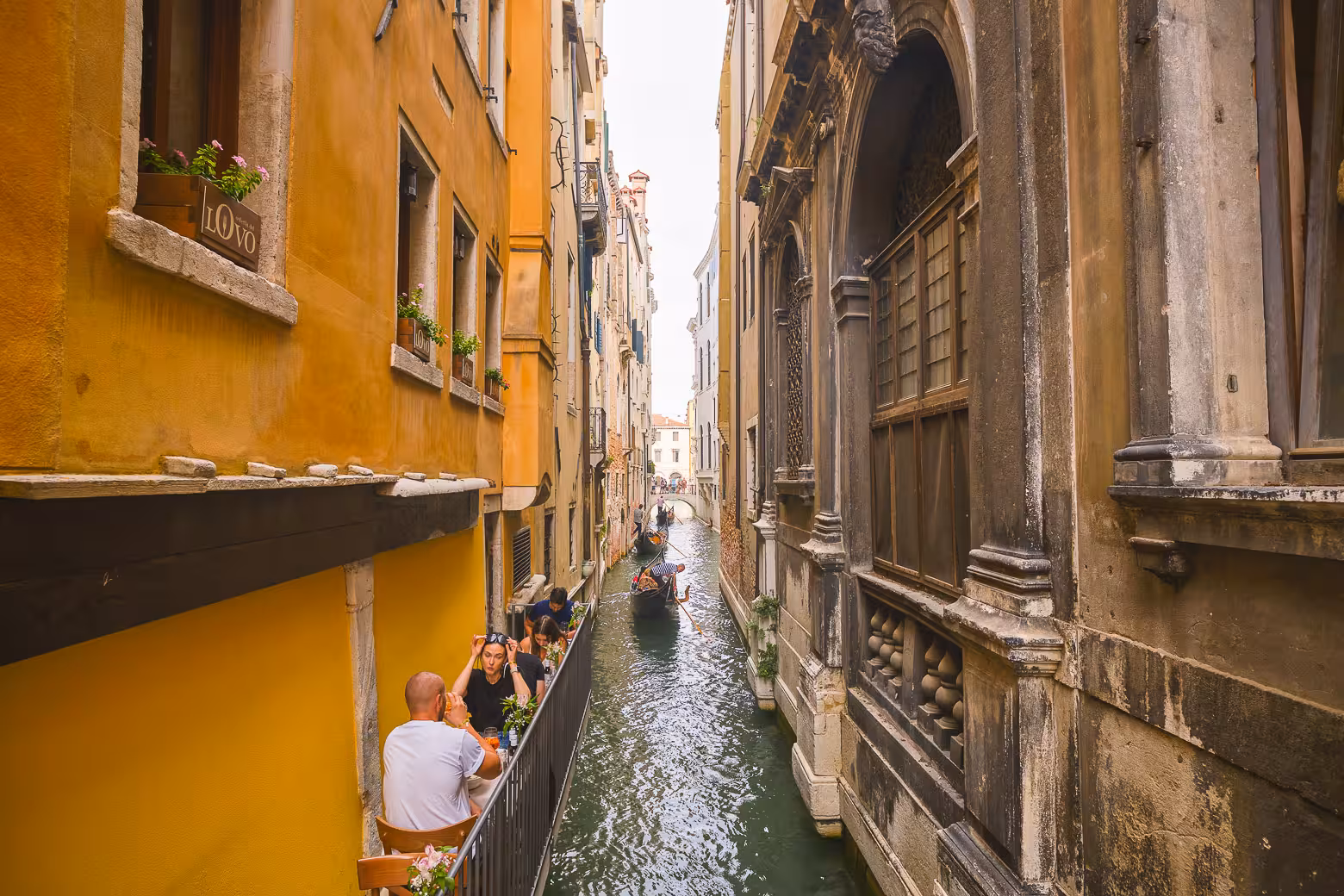 Narrow canal with gondola passing by charming diners at an outdoor cafe in Venice's historic district.