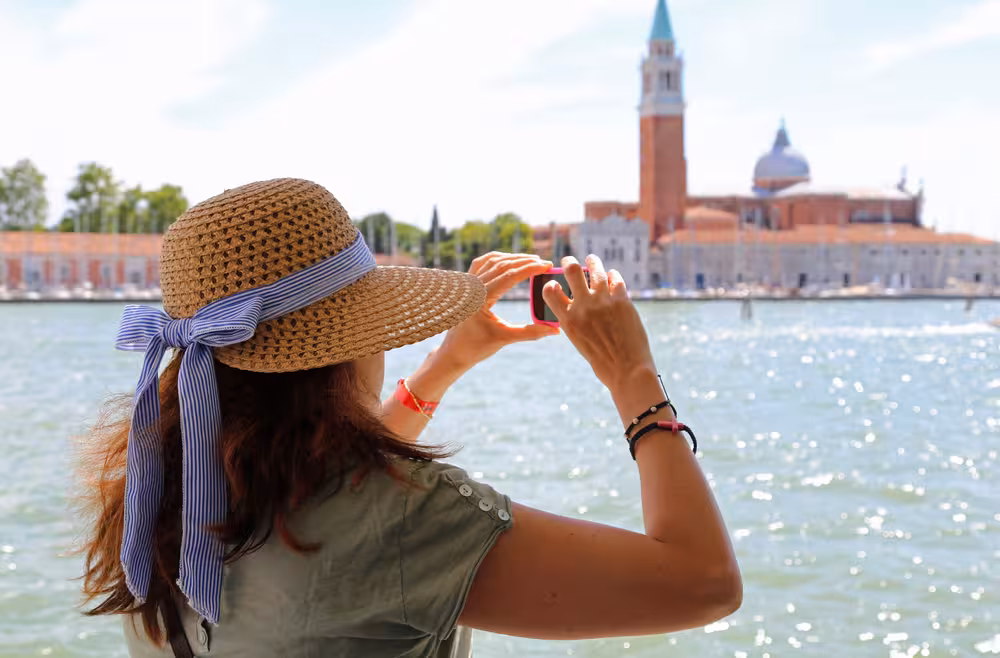 Woman in a sun hat capturing a photo of San Giorgio Maggiore's iconic bell tower across the shimmering water.