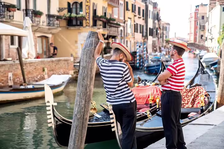 Two gondoliers in striped shirts chatting beside moored gondolas on a quiet Venice canal before a private gondola ride