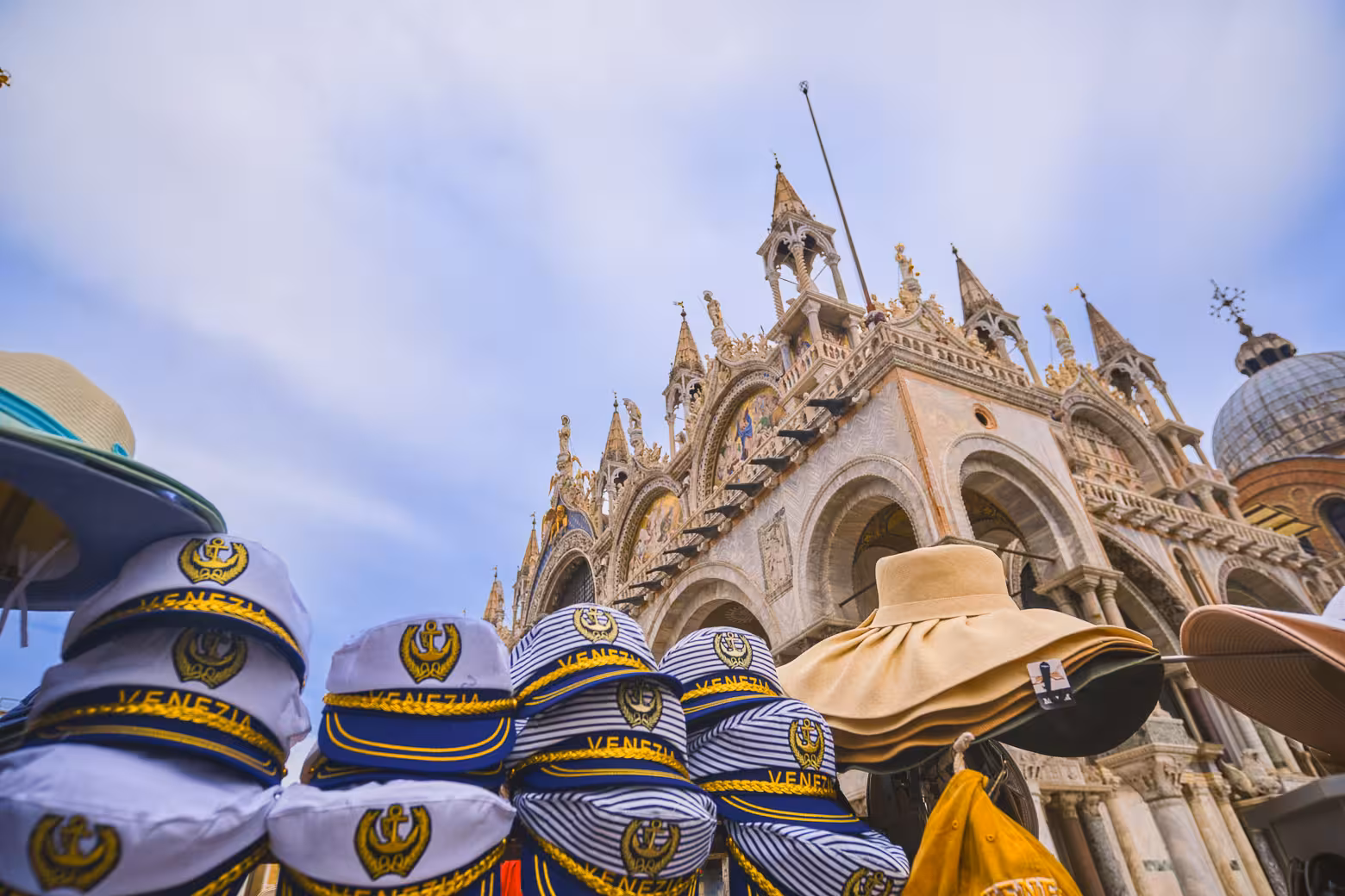 Venice souvenir hats with St. Mark's Basilica in the background on a sunny day, showcasing iconic Italian architecture.