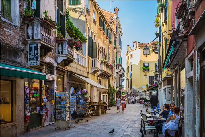 Charming Venice pedestrian street with colorful buildings, outdoor cafe seating, and a small bridge in the background