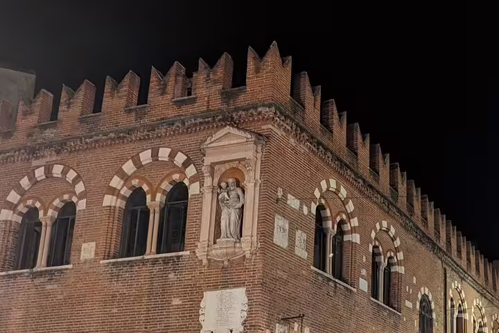 Night view of Verona’s medieval brick palace with crenellations and statue, featured on guided walking tour