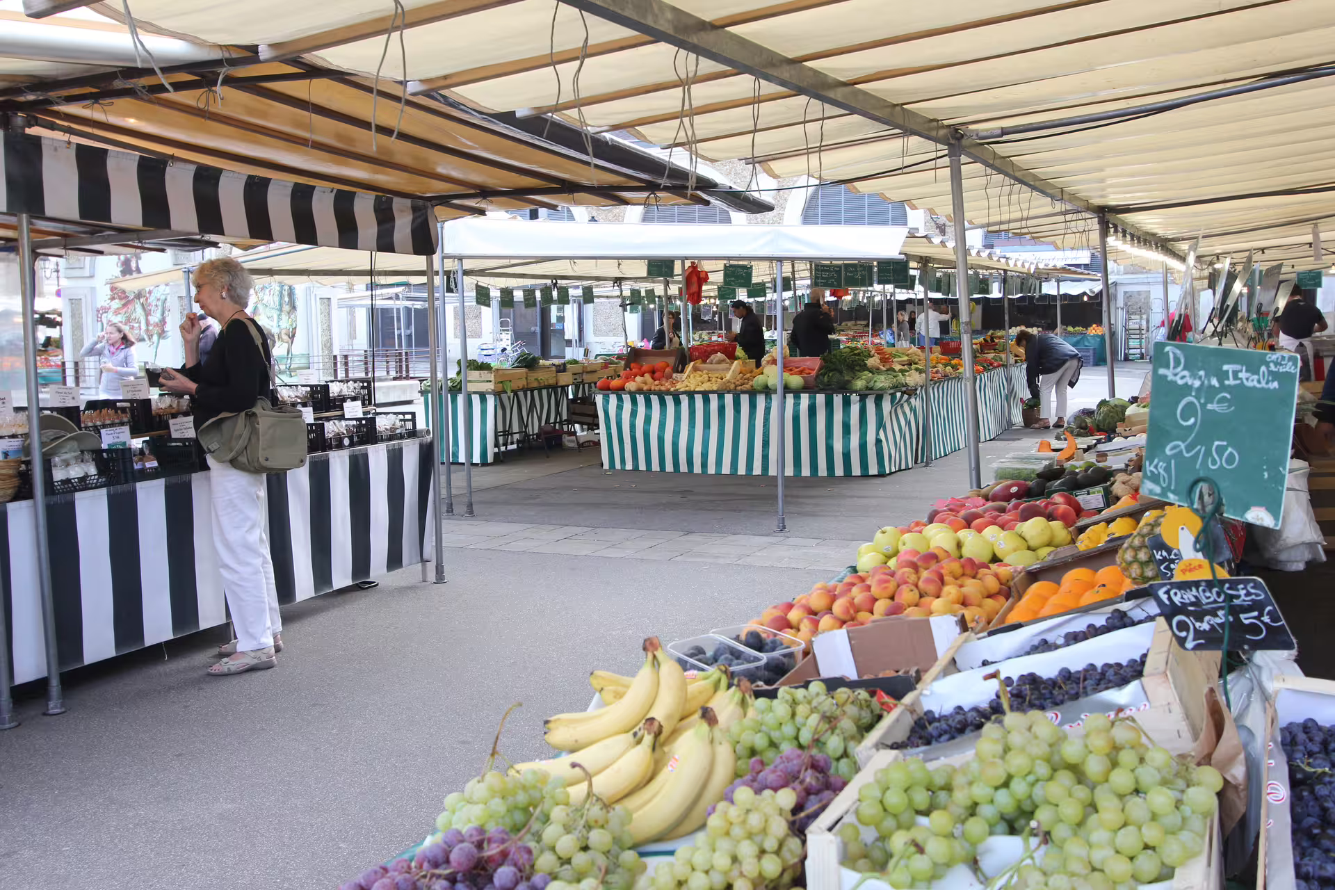Outdoor Versailles market stalls with fresh fruit, grapes, bananas, and produce under striped canopies