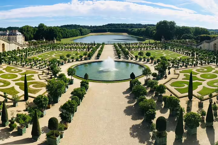 Aerial view of Palace of Versailles formal gardens with circular fountain, trimmed topiaries, and grand lake beyond