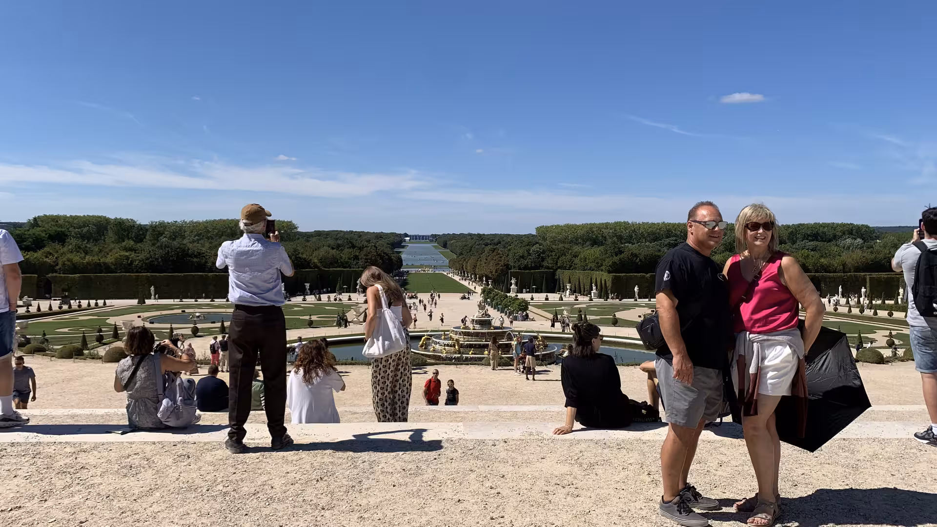 Tourists posing at the grand gardens of Palace of Versailles with fountain and tree-lined canal views