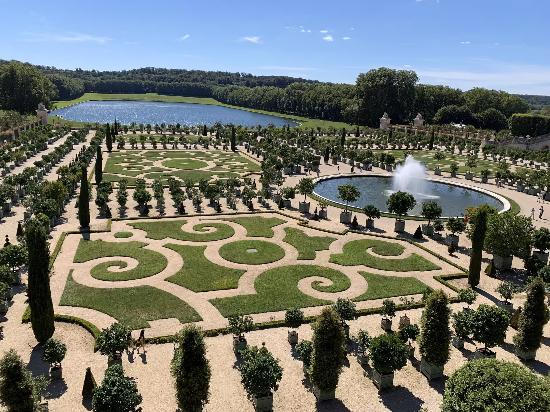 Aerial view of Versailles Palace ornate gardens with geometric parterre, fountain, and grand lake beyond