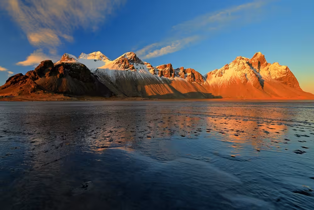 Snow-capped Vestrahorn mountain reflected in wet black sand beach at golden sunset, Iceland