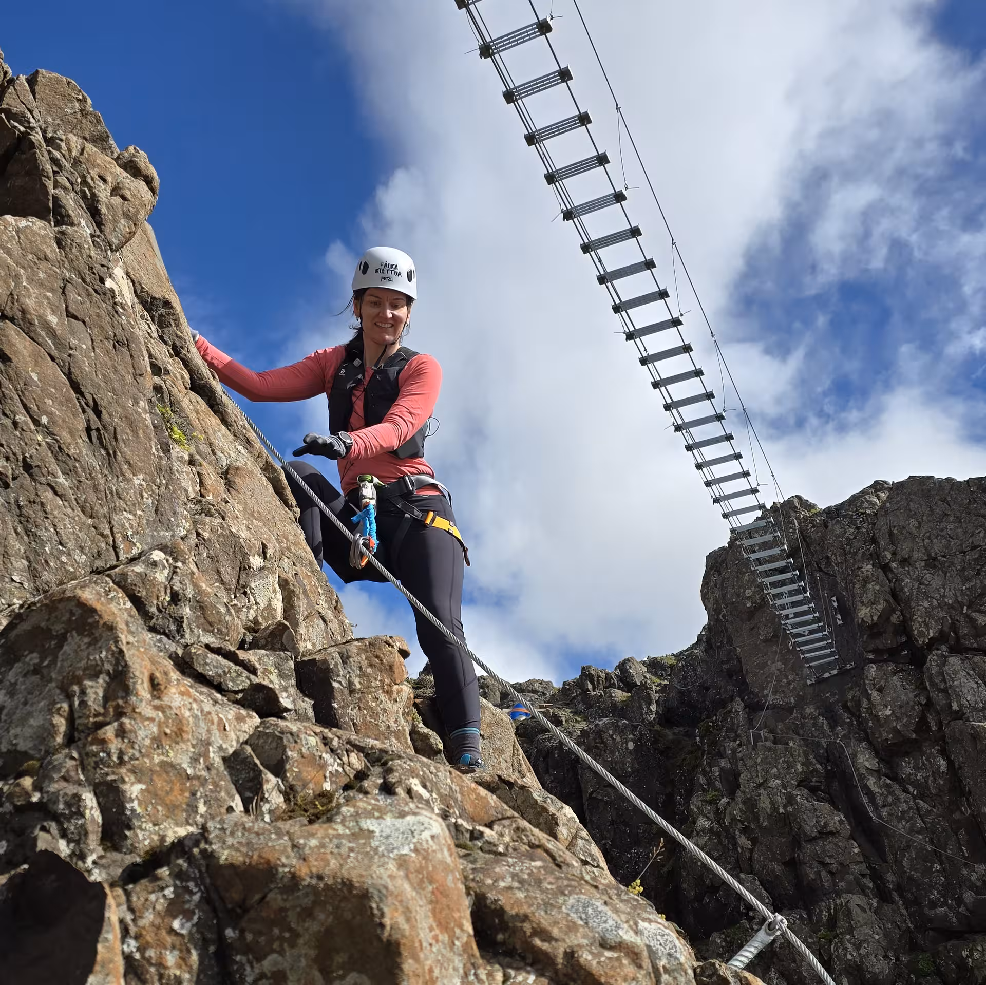 Woman in helmet and harness climbing rocky via ferrata route with suspension ladder at Falcon Rock Iceland