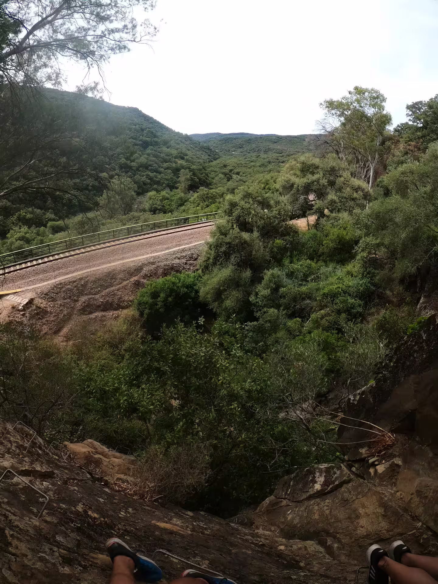 Adventurer climbs rocky Via Ferrata El Caimán trail with lush greenery and scenic railway view in Alcornocales Park.