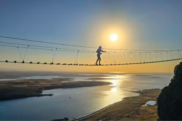 Person walking suspension bridge at Falcon Rock via ferrata with golden sunset and Reykjavik coastline below