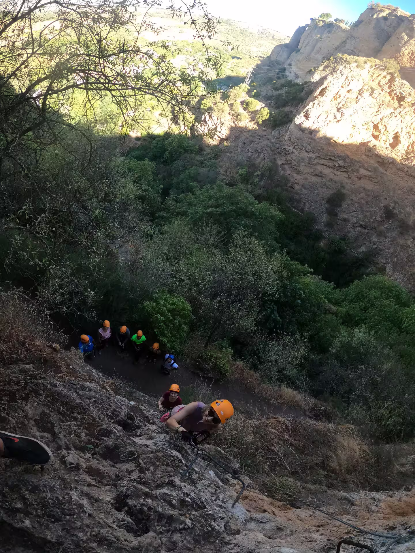 Climbers wearing helmets ascend the rugged cliff of Via Ferrata Tajo de Ronda, surrounded by lush greenery and dramatic rock formations.