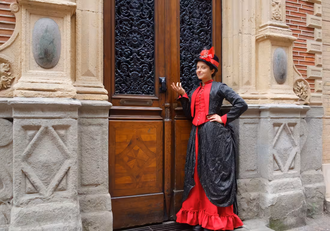 Guide in Victorian red and black dress posing at ornate wooden door in Toulouse Saint Georges district