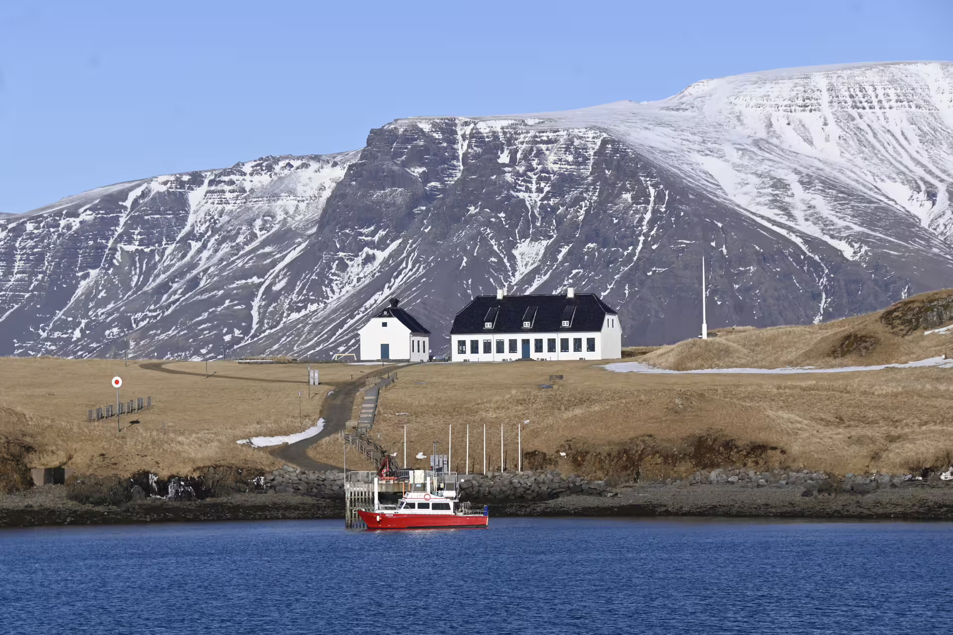 Red ferry boat docked at Videy Island pier with white building and snow-capped mountains in background